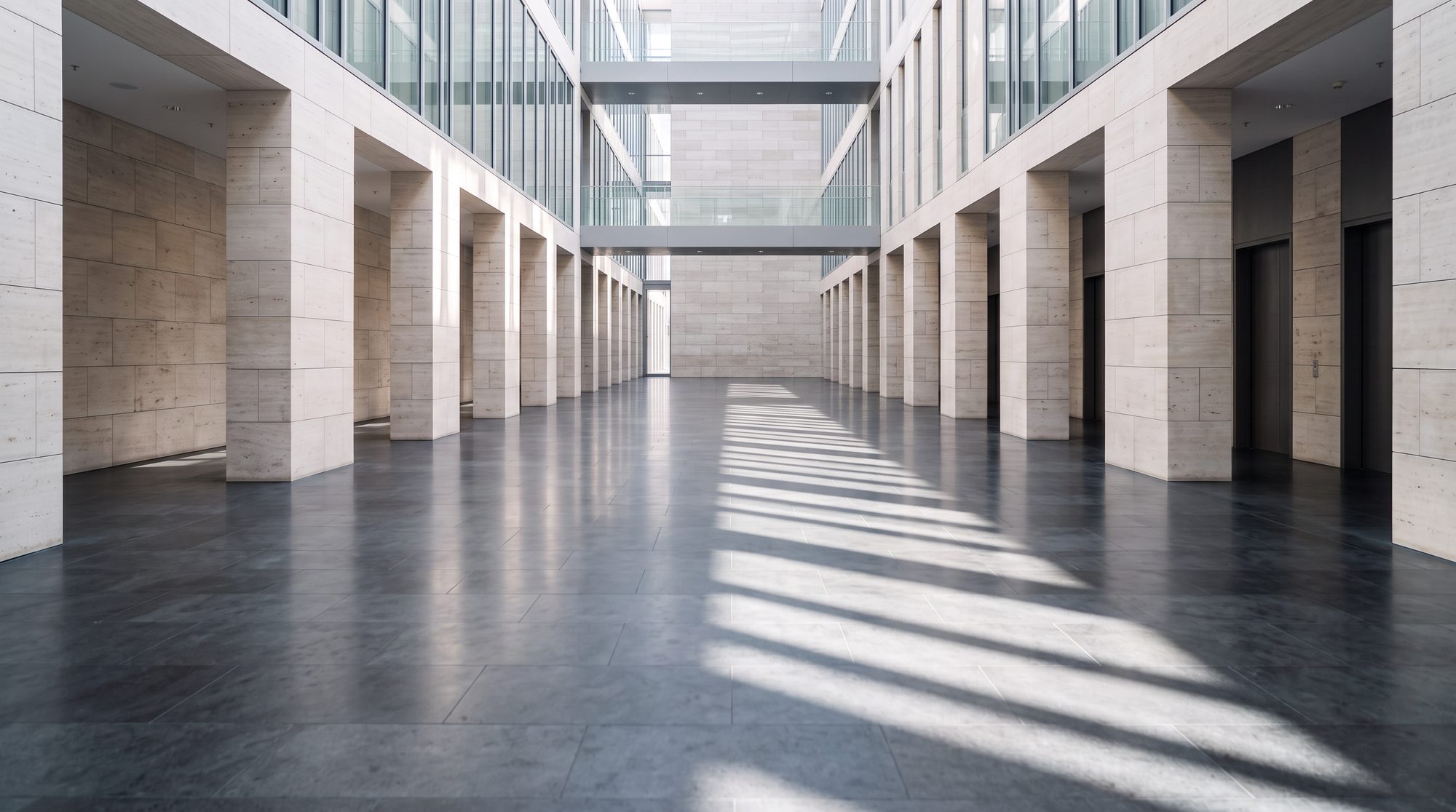 Galerie de pierre claire rythmée de piliers, vide, sous une lumière rasante de matin.